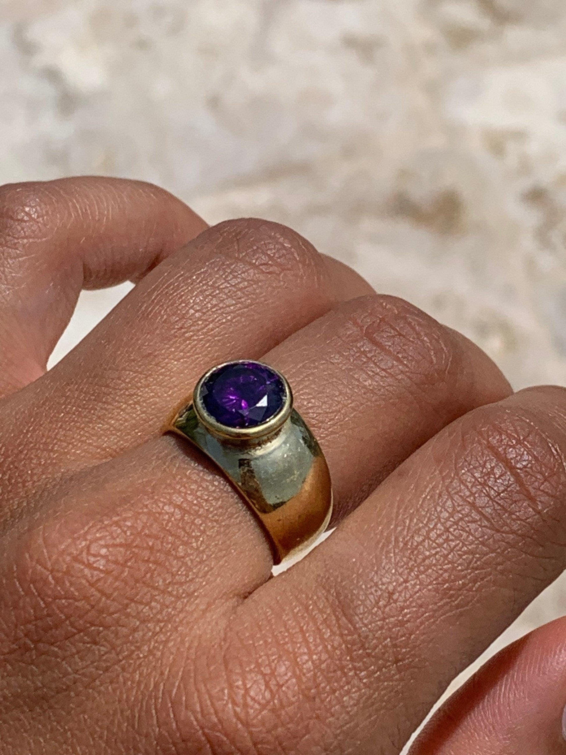 Könnte beinhalten: Ein goldener Ring mit einem großen, runden, violetten Edelstein, der in der Mitte eingesetzt ist. Der Ring ist dick und hat eine abgerundete Band.