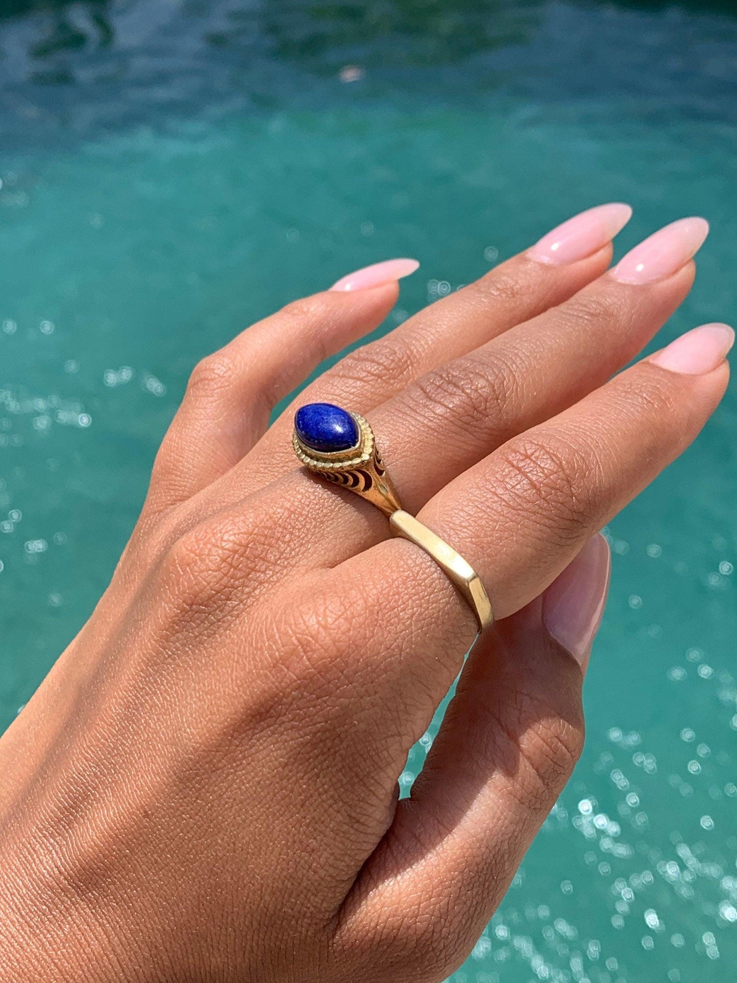 Könnte beinhalten: Ein goldener Ring mit einem blauen Edelstein, der in einem dekorativen Design gefasst ist. Der Ring wird an einem Finger mit einem zweiten goldenen Bandring getragen.