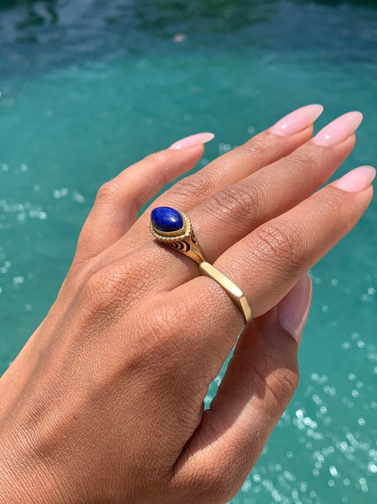 Könnte beinhalten: Ein goldener Ring mit einem blauen Edelstein, der in einem dekorativen Design gefasst ist. Der Ring wird an einem Finger mit einem zweiten goldenen Bandring getragen.